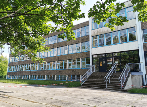 Teilansicht eines großen, dreistöckigen Gebäudes mit langen blau gerahmten Fensterreihen und dunkler Eingangstür zu der eine Treppe führt. Der Platz vor der Treppe ist mit rot-weißem Absperrband abgesperrt. 