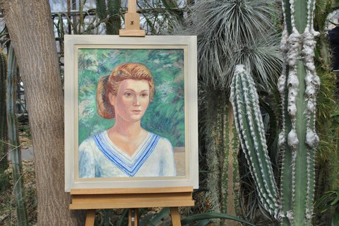 Ein Gemälde zeigt ein Portrait einer rothaarigen Frau. Hinter und neben dem Bild sind Kakteen und andere Pflanzen erkennbar.