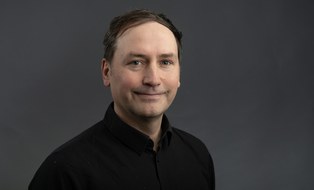 Ein Mann steht vor dunkelgrauem Hintergrund. Er trägt ein mit schwarzes Hemd und lächelt in die Kamera.