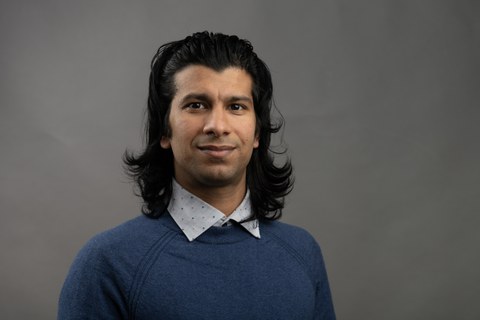 Ein Mann mit langem, schwarzem Haar und blauem Pullover steht vor einem dunkelgrauen Hintergrund und schaut in die Kamera.