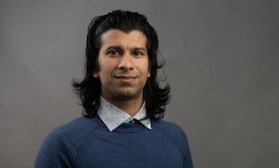 Ein Mann mit langem, schwarzem Haar und blauem Pullover steht vor einem dunkelgrauen Hintergrund und schaut in die Kamera.