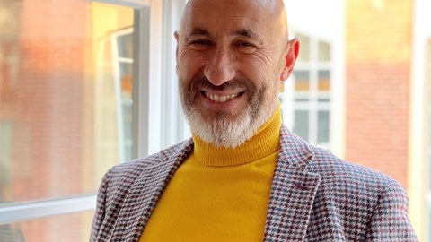 Herr Hurtado steht vor einem Fenster. Er trägt ein kariertes Sakko und ein gelbes Shirt. Er lacht in die Kamera.