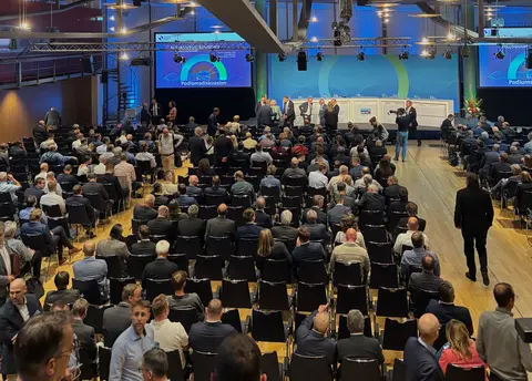 Zahlreiche Stuhlreihen in einer großen Halle mit Blickrichtung auf eine Bühne, flankiert von großen Monitoren. Auf der Bühne sind plätze für eine Podiumsdiskussion vorbereitet. Die Stuhlreihen sind teilweise besetzt. Einige Personen gehen durch die Halle. 