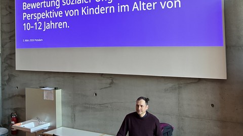 Doktor Matthias Ritter steht am Pult, vor ihm der aufgeklappte Laptop, die linke Hand in der Hosentasche, die rechte Hand stützt er auf dem Pult ab. Hinter ihm wird auf einem großen Bildschirm an der Wand der Titel seines Vortrags angezeigt.
