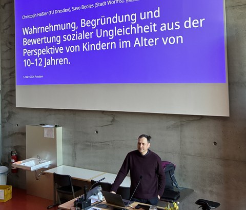 Doktor Matthias Ritter steht am Pult, vor ihm der aufgeklappte Laptop, die linke Hand in der Hosentasche, die rechte Hand stützt er auf dem Pult ab. Hinter ihm wird auf einem großen Bildschirm an der Wand der Titel seines Vortrags angezeigt.