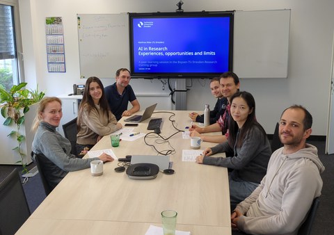 Eine Gruppe Menschen sitzt um einen Besprechungstisch. Alle lächeln in die Kamera. Im Hintergrund an der Wand ist ein großer Monitor mit einer Präsentation.