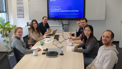 Eine Gruppe Menschen sitzt um einen Besprechungstisch. Alle lächeln in die Kamera. Im Hintergrund an der Wand ist ein großer Monitor mit einer Präsentation.