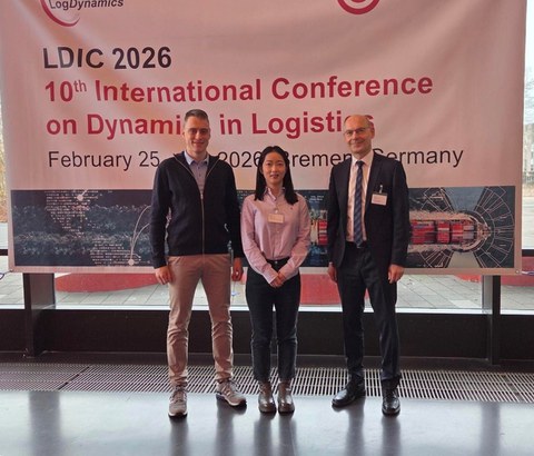 Die Kollegiatin Shiyu Guo steht mir ihrem Betreuer Professor Udo Buscher und einem weiteren Konferenzteilnehmer vor einem großen Banner mit der Aufschrift LDIC 2026 10th International Conference on Dynamics in Logistics 