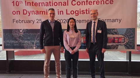 Die Kollegiatin Shiyu Guo steht mir ihrem Betreuer Professor Udo Buscher und einem weiteren Konferenzteilnehmer vor einem großen Banner mit der Aufschrift LDIC 2026 10th International Conference on Dynamics in Logistics 