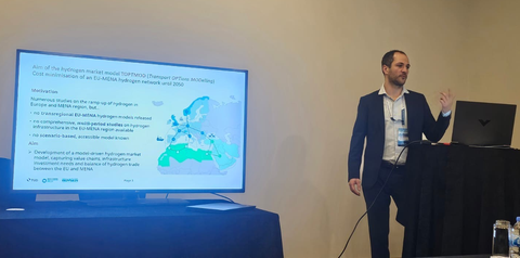 Jakob Baumgarten steht gestikulierend an einem Pult. Im Hintergrund ein großer Bildschirm mit einer Präsentation.
