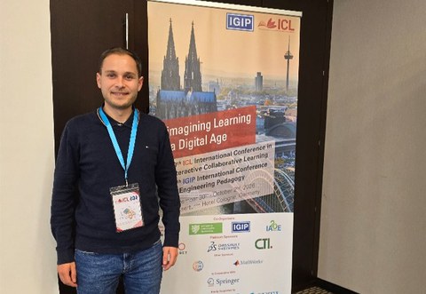 Kollegiat Jannik Amon steht neben einem Aufsteller mit einem Plakat zur Konferenz in Köln. Auf dem Plakat rechts von ihm ist der Kölner Dom zu sehen.