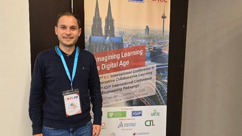Kollegiat Jannik Amon steht neben einem Aufsteller mit einem Plakat zur Konferenz in Köln. Auf dem Plakat rechts von ihm ist der Kölner Dom zu sehen.
