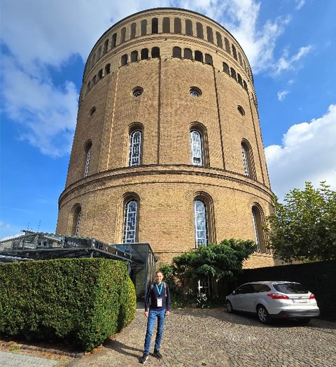 Ein Mann steht vor einem turmförmigen Gebäude aus braunem Stein mit großen Rundbogenfenstern.