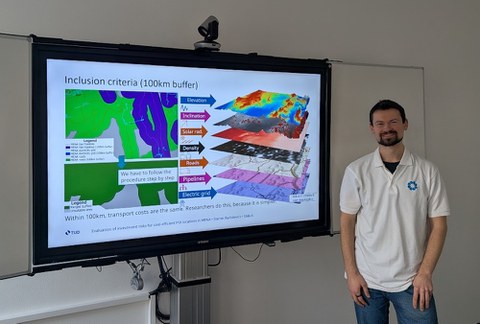 Kollegiat Siarhei Bartalevich steht lächelnd neben einem großen Monitor auf dem ein teil seiner Präsentation zu sehen ist.