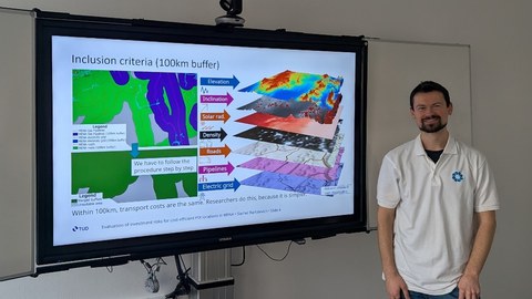 Kollegiat Siarhei Bartalevich steht lächelnd neben einem großen Monitor auf dem ein teil seiner Präsentation zu sehen ist.
