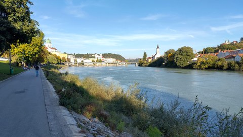 Links ein Weg, der am Ufer eines Flusses verläuft. Im Hintergrund sieht man in der Ferne die Stadt Passau.