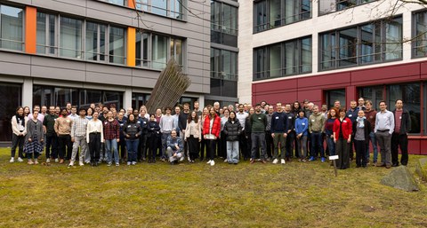 Die Teilnehmenden der Konferenz stehen im Freien auf einer Wiese, im Hintergrund ist ein mehrstöckiges Gebäude zu sehen.