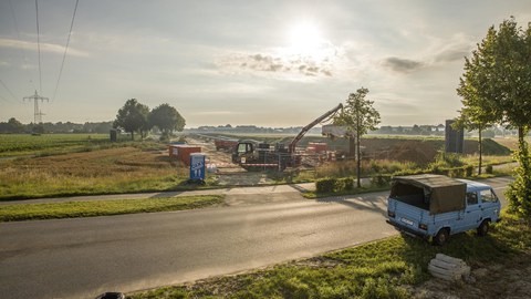 Im Vordergrund eine Landstraße mt einem kleinen Lieferwagen. Im Hintergrund ein Feld mit einem Baukran auf einer Baustelle.
