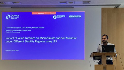 Ein Mann steht rechts an einem Pult, vor ihm ein Laptop, links hinter ihm wird eine Präsentation an die Wand projeziert.