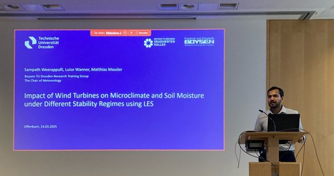 Ein Mann steht rechts an einem Pult, vor ihm ein Laptop, links hinter ihm wird eine Präsentation an die Wand projeziert.