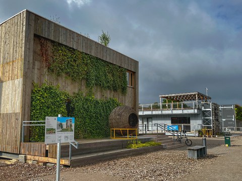 Ansicht des Hauses der Ungarn auf dem Gelände des Living Lab NRW, zweigeschossige Fassade mit Holzverschalung auf Sockel, davor Korbobjekt