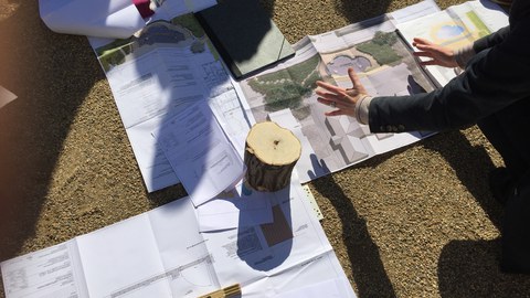 Foto zeigt auf dem Boden augebreitete Pläne, Skizzen und technischen Zeichnungen. Eine Lehrperson erklärt diese, während die Studenten darum verteilt stehen.