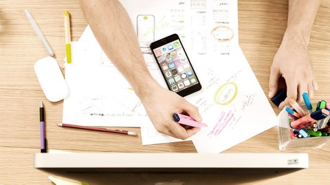 Foto auf einen Arbeitsplatz von oben gesehen mit Bildschirm, Handy, Papier und Stiften