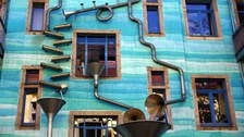 Das Foto zeigt den 'Hof der Elemente' in der Kunsthofpassage in der Dresdener Neustadt. An dem dargestellten Haus sind verschieden gebogene Rohre befestigt, durch die Regenwasser laufen kann.
