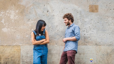 Eine junge Frau und ein junger Mann lehnen an einer Betonwand und lachen zusammen.