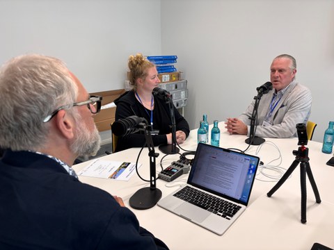 Drei Personen sitzen bei der Podcast-Aufnahme um Mikrophone.