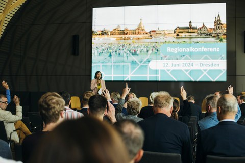 Im Vordergrund Publikum, im Hintergrund die Moderatorin der Konferenz auf der Bühne