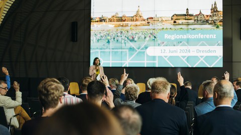 Im Vordergrund Publikum, im Hintergrund die Moderatorin der Konferenz auf der Bühne