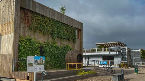 Ansicht des Hauses der Ungarn auf dem Gelände des Living Lab NRW, zweigeschossige Fassade mit Holzverschalung auf Sockel, davor Korbobjekt