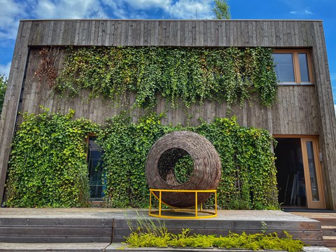 Ansicht des Hauses der Ungarn auf dem Gelände des Living Lab NRW, zweigeschossige Fassade mit Holzverschalung auf Sockel, davor Korbobjekt