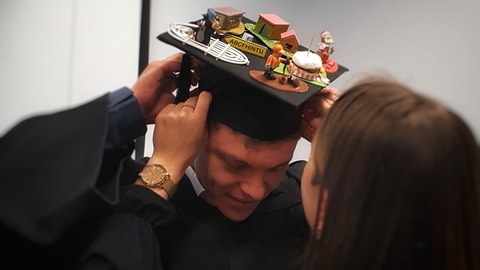 Der frisch promovierte Robin Zeh erhält den Doktorhut aufgesetzt. Auf dem Hut sind Modelle in Bezug auf seine Arbeit abgebildet.