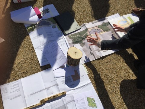 Foto zeigt auf dem Boden augebreitete Pläne, Skizzen und technischen Zeichnungen. Eine Lehrperson erklärt diese, während die Studenten darum verteilt stehen.