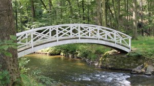 Foto zeigt eine weiße Holzbrücke im Seifersdorfer Tal, die einen Fluss überquert. 