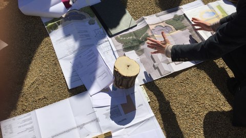 Foto zeigt auf dem Boden augebreitete Pläne, Skizzen und technischen Zeichnungen. Eine Person erklärt diese, während die Studenten darum verteilt stehen.