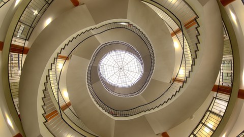 Treppenaufgang im Hülße-Bau der TU Dresden. Das Bild wurde von unten aufgenommen. Den Mittelpunkt bildet das Dachfenster, dessen Konstruktion wie ein Spinnennetz aussieht. Von unten betrachtet läuft die Treppe spiralförmig nach oben. 