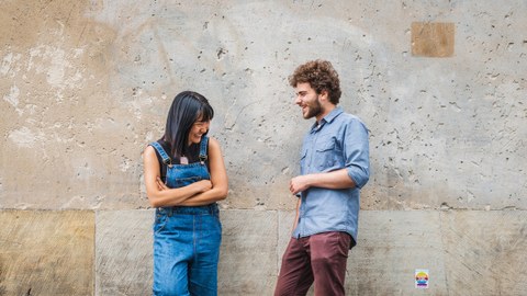 The picture shows a young woman and an young man leaning against a wall and laughing together.