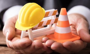 The photo shows two hands holding small construction figures. You can see a helmet, a traffic cone and a barrier.