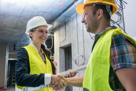 The photo shows a man and a woman on a construction site who are shaking hands. They are wearing helmets and yellow high-visibility vests.