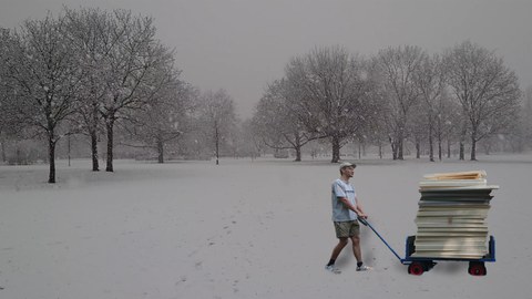 Das Bild zeigt einen Handwagen mit Modellbaupappen welcher von einem Studierendem durch den Schnee gezogen wird. 