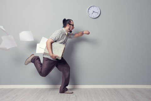 In the photo you see a man. He runs and loses a lot of papers. The man looks at his watch and runs past a wall on which another watch is hanging.