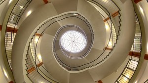 Treppenaufgang im Hülße-Bau der TU Dresden. Das Bild wurde von unten aufgenommen. Den Mittelpunkt bildet das Dachfenster, dessen Konstruktion wie ein Spinnennetz aussieht. Von unten betrachtet läuft die Treppe spiralförmig nach oben. 