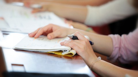 Foto einer Hand, die einen Stift hält und auf einem Hefter liegt