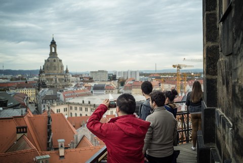 Eine Gruppe Studierender bei einer Stadtbesichtigung auf dem Hausmannsturm in Dresden. Sie blicken auf die Stadt herab. Ein junger Mann in roter Jacke macht ein Foto von der Frauenkirche. 