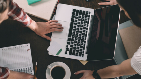 Draufsicht auf einen Tisch wo drei LEute arbeiten, mit Laptop, Notizen und Kaffe