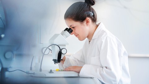 Eine Wissenschaftlerin in einem weißen Kittel schaut in ein Mikroskop. 