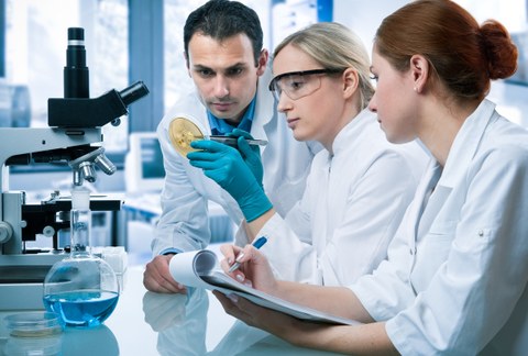 Das Foto zeigt drei Personen in weißen Laborkitteln. Sie sitzen vor einem Mikroskop. Eine der Personen hält eine Petrischale mit Bakterien in der Hand. Eine andere Person macht Notizen auf einem Block.